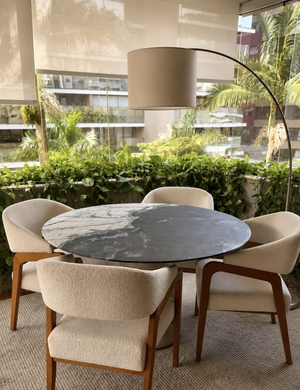 Mesa de jantar redonda em varanda com poltronas claras e vegetação.